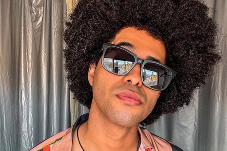 Homem em primeiro plano, de óculos escuros, faz pose para a câmera com cortina ao fundo