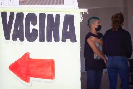 Placa escrito vacina com seta vermelha e menino de preto e máscara ao fundo
