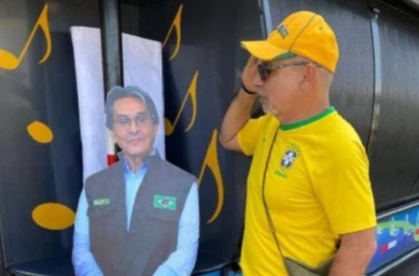 Na imagem colorida, um homem está posicionado no centro. Ele é careca e está com as mãos levantadas