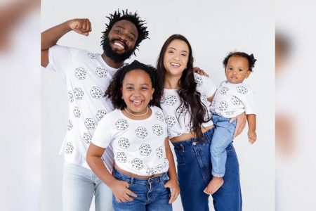 Na imagem colorida, quatro pessoas estão centralizadas no meio. Elas usam camiseta branca estampada com dados e jeans. Estão aglomeradas e sorriem para a camera
