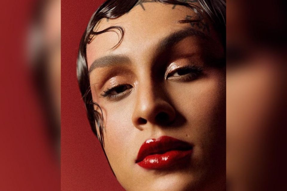 Na imagem colorida, uma mulher está posicionada no centro. Ela tem cabelos escuros e curtos, tem olhos castanhos e olha seriamente para a câmera