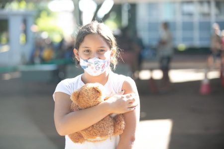 Menina com cabelo amarrado, máscara no rosto segurando ursinho de pelúcia marrom no braço