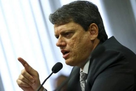 Na imagem colorida, um homem está posicionado no centro. Ele tem cabelos curtos, pretos e olha seriamente para a camera