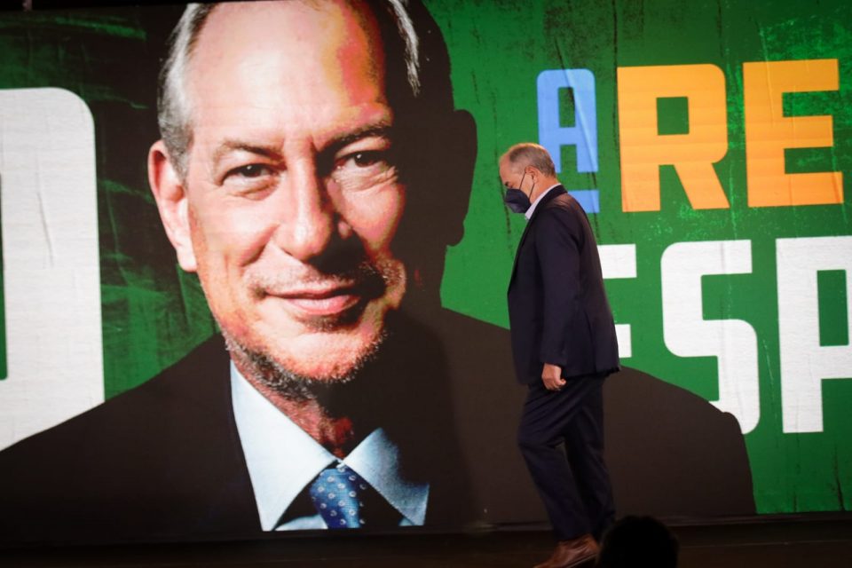 Ciro Gomes de perfil, andando de máscara, fotografado sob painel com sua própria foto sorrindo - Metrópoles