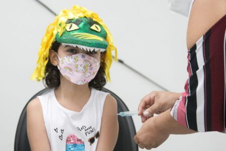 Menina com chapéu de jacaré na cabeça e máscara sendo vacinada