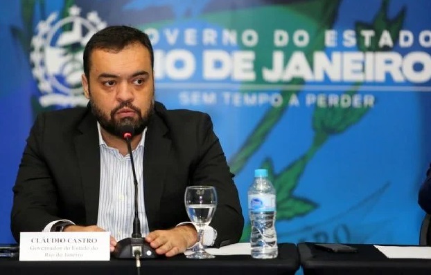 Na imagem colorida, um homem está posicionado no centro. Ele tem cabelos curtos, escuros, usa camiseta clara, terno escuro e olha seriamente para a câmera
