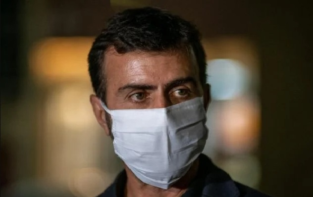 Na imagem colorida, um homem está posicionado no centro. Ele tem cabelos curtos, escuros, usa camiseta clara, terno escuro, mascara branca e olha seriamente para a camera