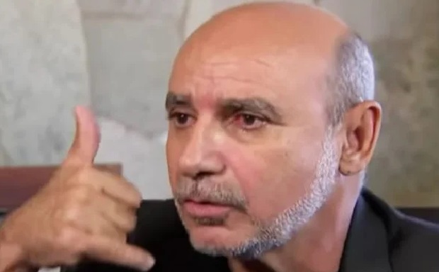 Na imagem colorida, um homem está posicionado no centro. Ele tem cabelos curtos, pele clara e olha seriamente para a frente