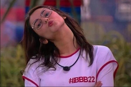 Eslôvenia veste roupa clara com o nome do programa escrito do lado esquerdo superior da roupa. Ela tem cabelos escuros, usa óculos e está mandando beijos- Metrópoles
