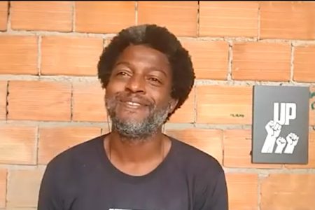 Homem negro de camisa preta, em primeiro plano, com uma parede de tijolos descobertos ao fundo.