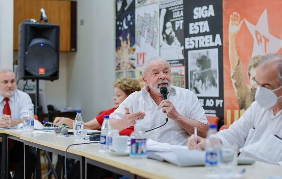 Na foto, sentados numa mesa longa, estão o senador Jean Paul Prates, a ex-presidente Dilma Rousseff, o ex-presidente Lula e o ex-senador Aloísio Mercadante, todos do PT. Lula discursa num microfone, olhando para frente, numa sala com cartazes do PT - Metrópoles