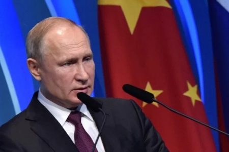 Sob fundo azul com parte da bandeira da China, o presidente russo Vladimir Putin fala em evento. Dois microfones estão posicionados a sua frente - Metrópoles