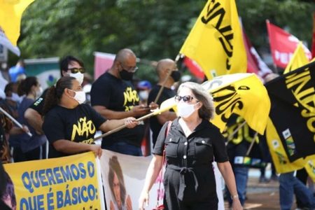 Pessoas estão segurando bandeiras e cartazes na cor amarela. Pessoas se aglomeram em um espaço público fazendo alguma espécie de manifestação e usam roupas pretas com as mesmas nomenclaturas com estampas. As blusas são pretas e as frases nelas têm a cor amarela