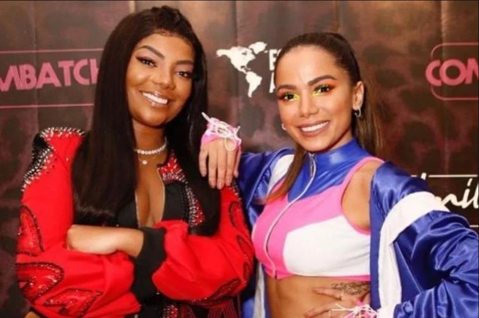 Cantora Ludmilla posando ao lado da cantora Anitta. Ludmilla usa casaco vermelho com preto e Anitta usa top branco com rosa e roxo. Alas sorriem para a câmera - Metrópoles