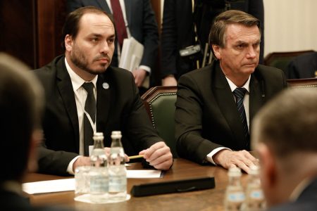 Vereador do Rio Carlos Bolsonaro acompanhou o pai, Jair Bolsonaro, na viagem à Rússia. Eles estão sentado numa mesa com outras pessoas, ambos de terno, sem máscara e com expressão séria - Metrópoles