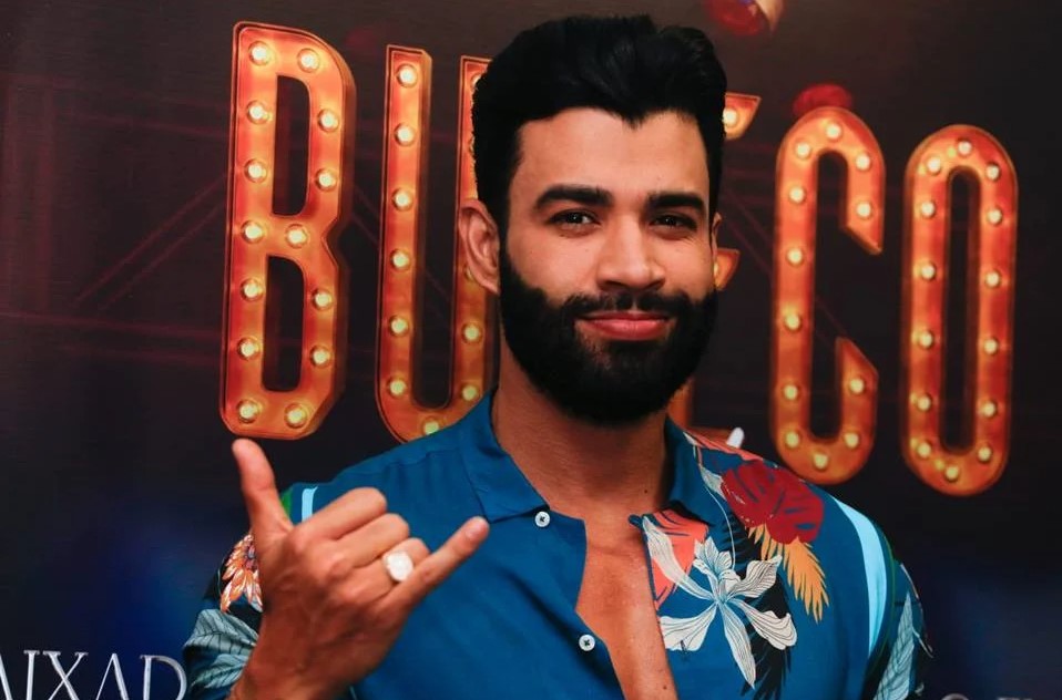 Gusttavo Lima cantor sertanejo em show. Ele usa camiseta azul estampada e tem cabelos pretos e barba preta