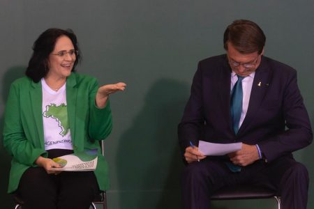 Em evento no Planalto do projeto "Brasil para Todos", a ministra Damares Alves sorri e faz um gesto ao público enquanto Bolsonaro, de óculos, lê um texto apoiado em sua perna - Metrópoles