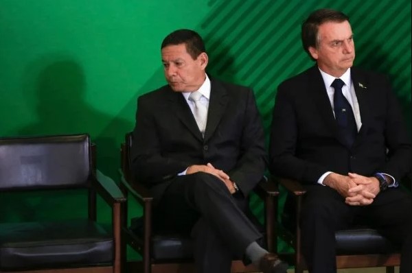 Bolsonaro e Mourão olham para lados opostos em cerimônia. Eles vestem terno e gravata e tem cabelos escuros - Metrópoles