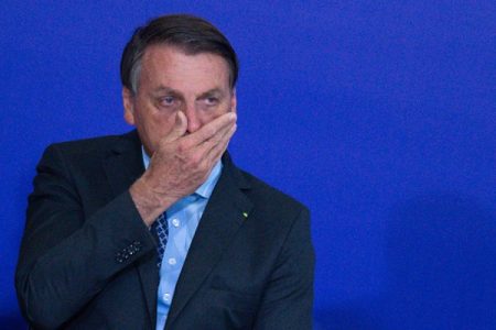 Bolsonaro tampa a boca com uma das mãos. Ele veste terno preto e camisa azul-Metrópoles