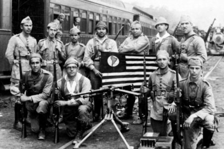 Foto em preto e branco. Participantes da Revolução Constitucionalista de 1932 posam juntos para foto com bandeira de São Paulo
