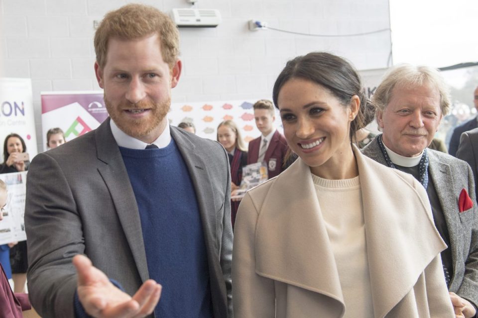 Foto colorida. Príncipe Harry com blazer e casaco azul. Meghan Markle com roupas beges