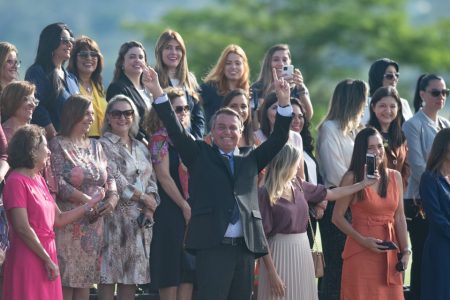 O presidente Jair Bolsonaro acena com as duas mãos levantadas, sorrindo, na frente de diversas mulheres