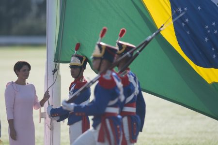 A primeira-dama Michelle Bolsonaro, com um vestido rosa, hasteia a bandeira do Brasil, enquanto, na frente dela, soltados aparecem com armas, chapéu e com trajes militares nas cores branca, azul e vermelha