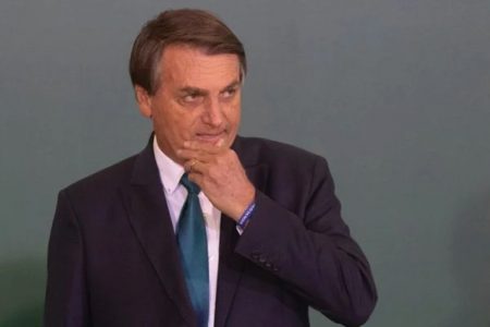 O presidente Bolsonaro em cerimônia no Planalto. Ele leva a mão ao rosto e olha para frente, usando terno - Metrópoles