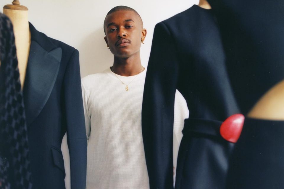 Homem negro e careca encostado na parede. Seu nome é Maximilian Davis e ele é estilista da marca Salvatore Ferragamo. Ele usa uma camiseta branca e posa ao lado de manequins usando blazers preto e azul.