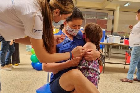Criança sentada em colo de mulher, com a cabeça para trás enquanto mulher aplica vacina em seu braço