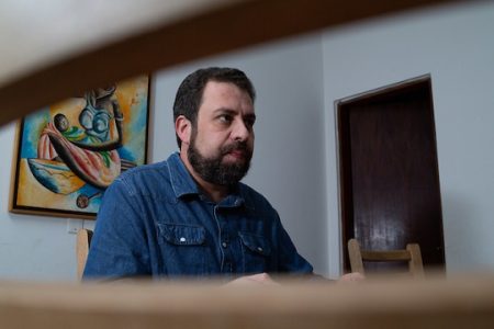 Imagem colorida mostra Guilherme Boulos de camisa azul marinho, sentado e com um quadro ao fundo - Metrópoles