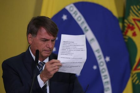O presidente Bolsonaro lê discurso no Planalto no lançamento do Programa Renda e Oportunidade. Ele usa terno e segura folha de papel, frente ao microfone com a bandeira do Brasil ao fundo - Metrópoles