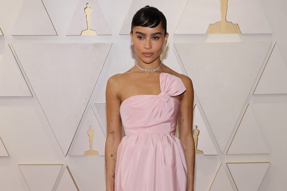 A atriz Zoë Kravitz no tapete vermelho do Oscar 2022. Ela é uma mulher negra, com cabelo presto e uma franja curta, e usa um vestido rosa claro da marca Yves Saint Laurent