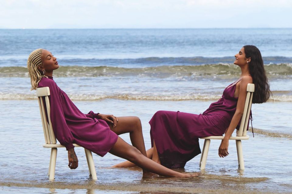 Modelos usando looks roxos, sentadas em cadeiras na beira do mar