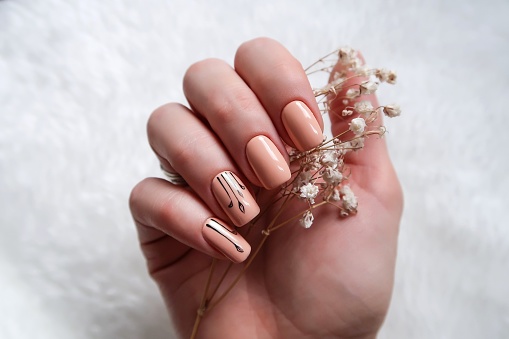 Mão com unhas pintadas de bege, algumas com desenhos em preto, segura um ramo de flores secas sob fundo branco - Metrópoles