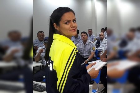 Mulher com casaco amarelo e azul e homens de farda sentados ao fundo