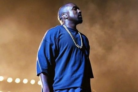 Foto colorida do rapper Kanye West em cima do palco com camiseta azul e corrente olhando para o público - Metrópoles