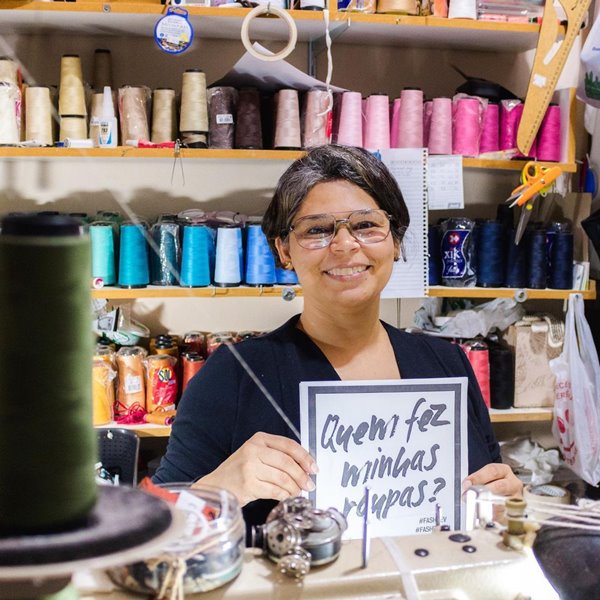 Trabalho Fashion Revolution Mulher morena de cabelo liso curto e de óculos de grau. Ela segura uma placa com os dizeres "quem faz as minhas roupas?" e está sentada em uma mesa com máquina de costura. No fundo é possível ver rolos de linha de diferentes cores - Metrópoles