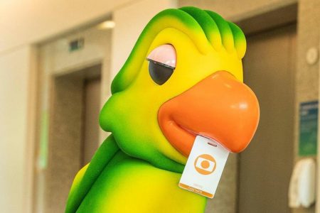 Boneco Louro José, mascote do programa Mais Você, segura crachá da Globo com o bico - Metrópoles