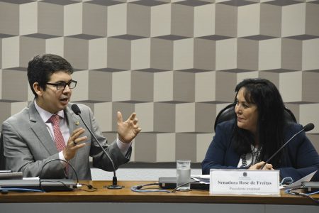 Os senadores Randolfe Rodrigues e Rose de Freitas sentados lado a lado na mesa diretora de uma comissão do Senado. Randolfe fala ao microfone enquanto Rose, sorrindo, o observa - Metrópoles