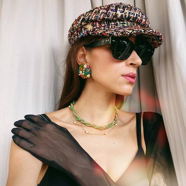 Mulher posa usando óculos escuros, boina de tweed, brincos redondos com pedras coloridas e luvas pretas