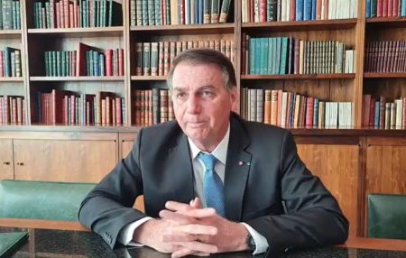 O presidente Bolsonaro durante fala em gabinete presidencial para vídeo. Ele está sentado e atrás é possível ver uma biblioteca cheia de livros nas estantes - Metrópoles