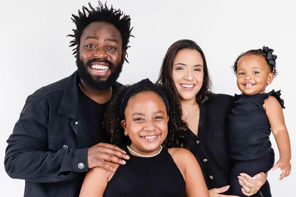 Família do brother do BBB22 Douglas Silva. Na foto, ele sorri com as duas filhas e a mulher, Carol Samarão, todos usando preto - Metrópoles