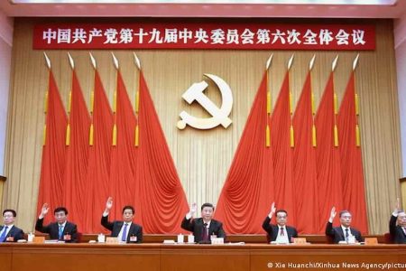 Salão principal da sede do Partido Comunista Chinês. Sete homens, incluindo o presidente chinês Xi Jin Ping, erguem as mãos em votação, e ao fundo é possível ver bandeiras vermelhas, escritos em ideogramas e o símbolo do comunismo - Metrópoles