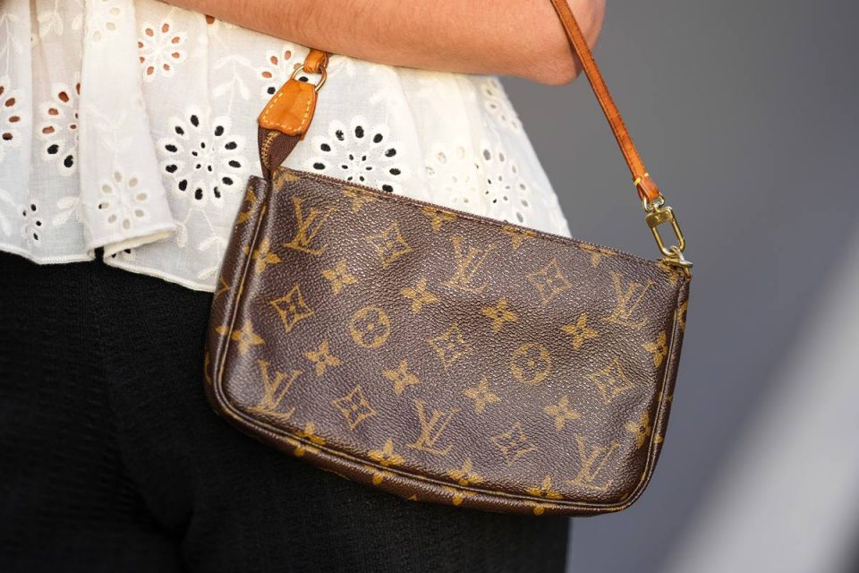 Mulher branca usando um top branco com babados/recortados, um padrão de impressão monograma marrom brilhante em uma bolsa de ombro de lona revestida da Louis Vuitton, calças pretas de pernas largas, durante uma sessão de fotos de moda de estilo de rua.