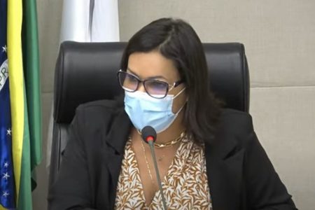 Mulher de máscara, óculos e blazer preto sentada em cadeira preta e falando ao microfone