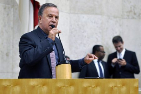 Camilo Cristófaro, homem branco de cabelos grisalhos, terno escuro, camisa escura e gravata lilás, discursa em microfone posicionado em cima de bancada - Metrópoles