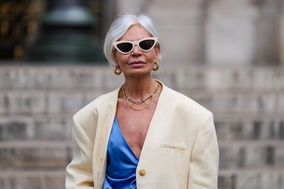 Mulher idosa branca, com cabelos curtos e também brancos, com roupa estilosa. Ela usa um óculos de sol vintage bege, um colar de pérolas multicoloridas, um colar de corrente de ouro, um blazer largo branco, um colete de seda azul brilhante com decote em V abotoado.