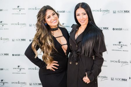 As irmãs Simone e Simaria, cantoras de sertanejo, tirando foto em um fundo de divulgação da turnê da dupla. Elas usam vestidos pretos justos e possuem a pele morena e os cabelos escuros longos.