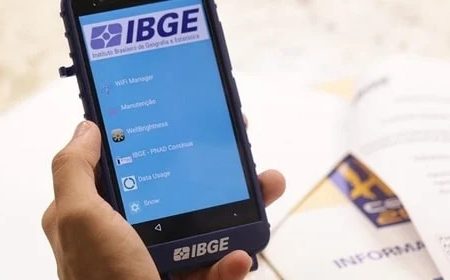 Mão segura aparelho do IBGE ligado sob mesa com papéis espalhados - Metrópoles